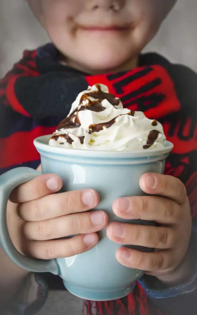 A childs hands hold a blue mug filled with creamy croc pot hot chocooate topped with a mound of swirly whip cream.