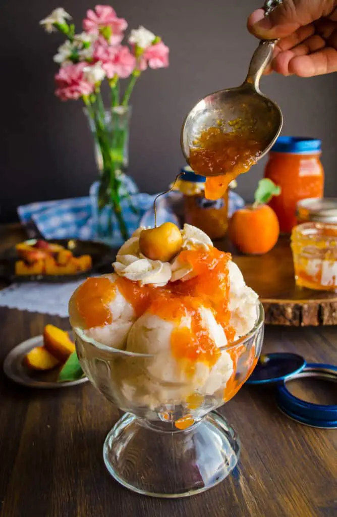 Homemade Vanilla Bean Peach Jam is drizzled onto a vanilla ice cream sundae with whipped cream and cherry on top.