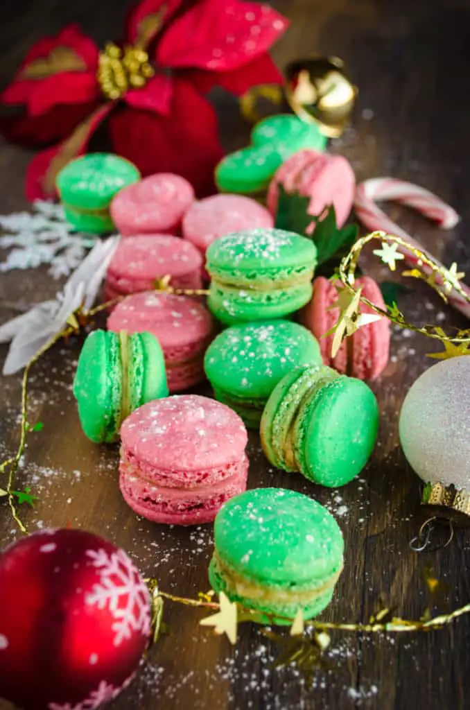 A closeup of red and green Christmas Macarons surrounded with Christmas Decorations and poinsetta leaves.