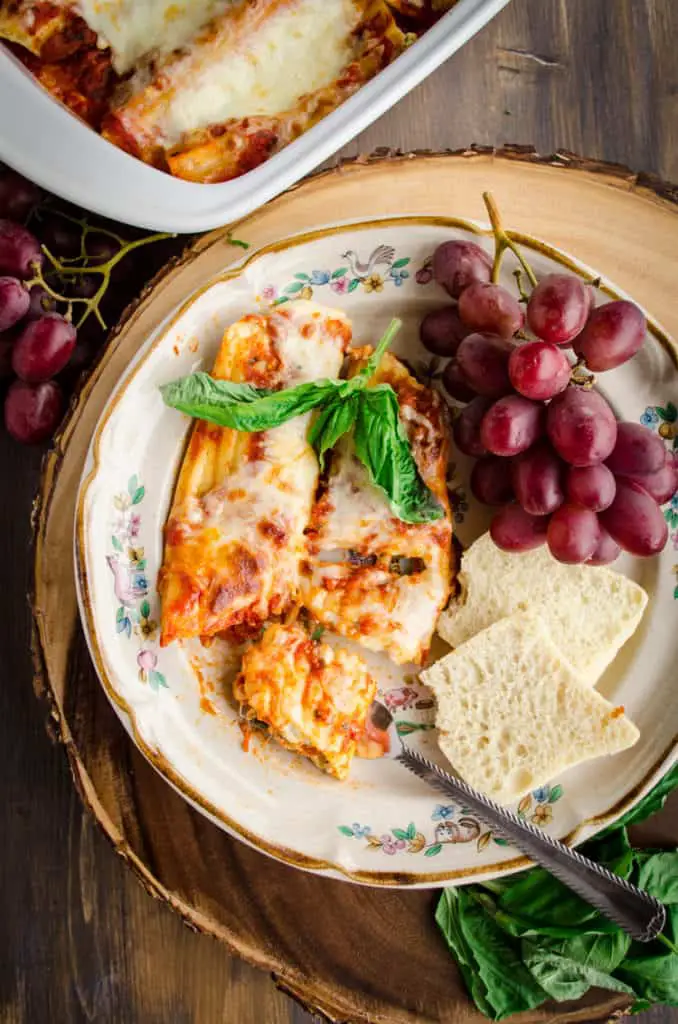a serving of Meatless Monday Cheese Stuffed Manicotti sits on a plate surrounded by red grapes, sliced baguettes, and fresh basil.