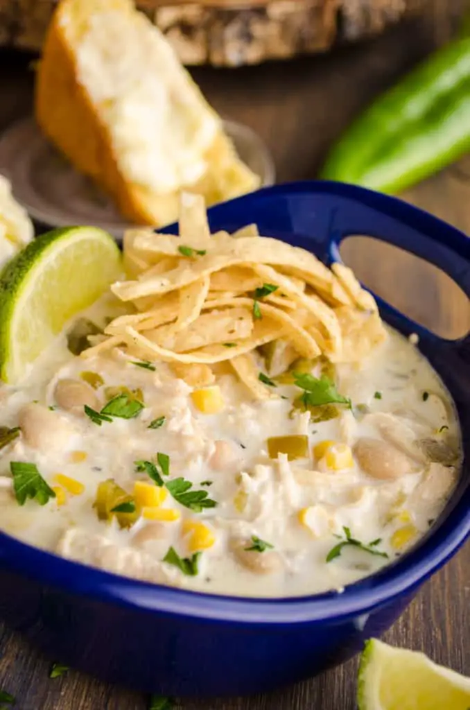A blue ceramic bowl of Creamy White Chicken Chili is garnished with tortilla strips, a lime wedge, and chopped cilantro.