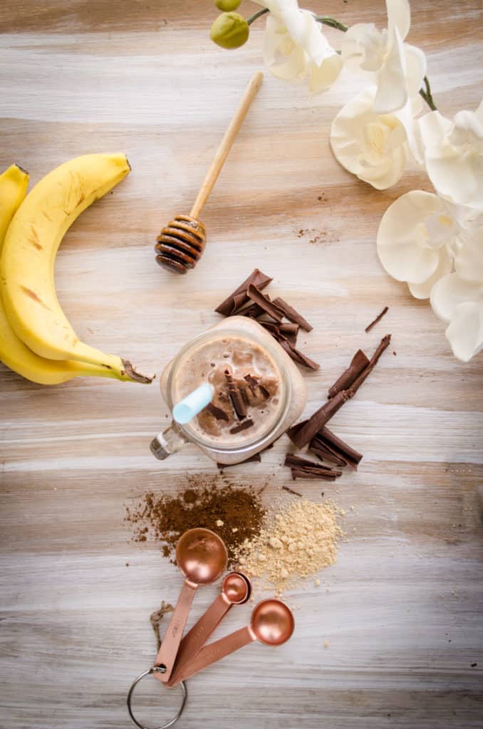 A top down view of a Peanut Butter Chocolate Banana Smoothie surrounded by bananas, measuring spoons, a honey server and vanilla flowers.