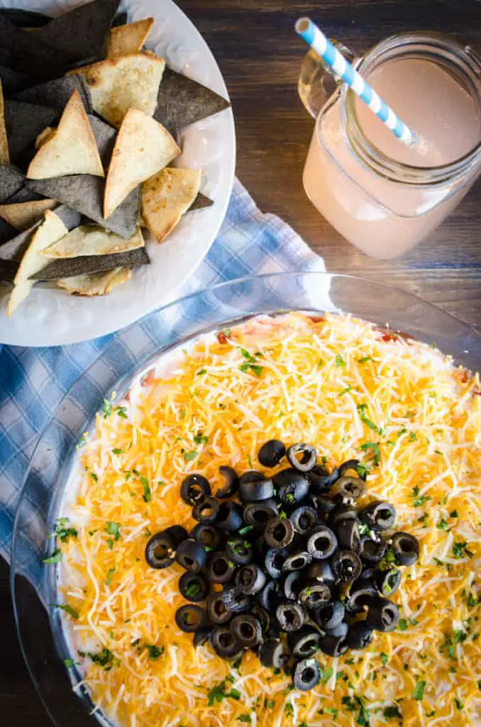 A top down view of a pie plate full of Spicy Chipotle Seven Layer Dip topped with black olives sits next to baked tortilla chips and a soda.