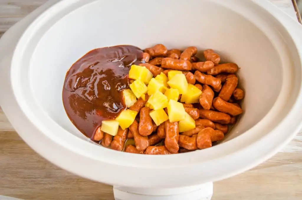 A slow cooker is filled with Lit'l smokies, pineapple chunks and barbecue sauce ready to cook into Slow Cooker Hawaiian Little Smokies - The Goldilocks Kitchen