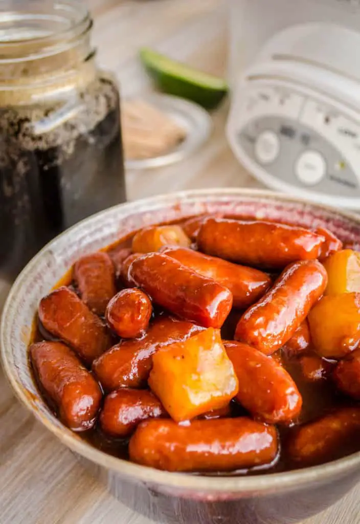 A bowl of Slow Cooker Hawaiian Little Smokies with chunks of pineapple in it sits in front of a slow cooker - The Goldilocks Kitchen