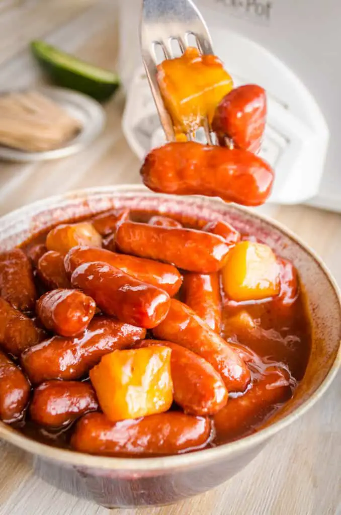 A close up of a bowl full of Slow Cooker Hawaiian Little Smokies with a fork holding a chunk of pineapple and a couple little smokies on the end - The Goldilocks Kitchen