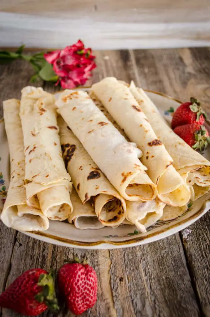 A plate filled with rolled Norwegian Lefse sits with a few strawberries and some red flowers. - The Goldilocks Kitchen