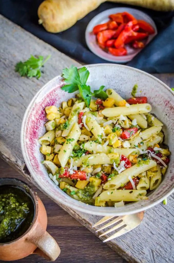 A bowl of Pesto Pasta with Roasted Root Veggies sits on a wooden plank, surrounded by fresh ingredients.