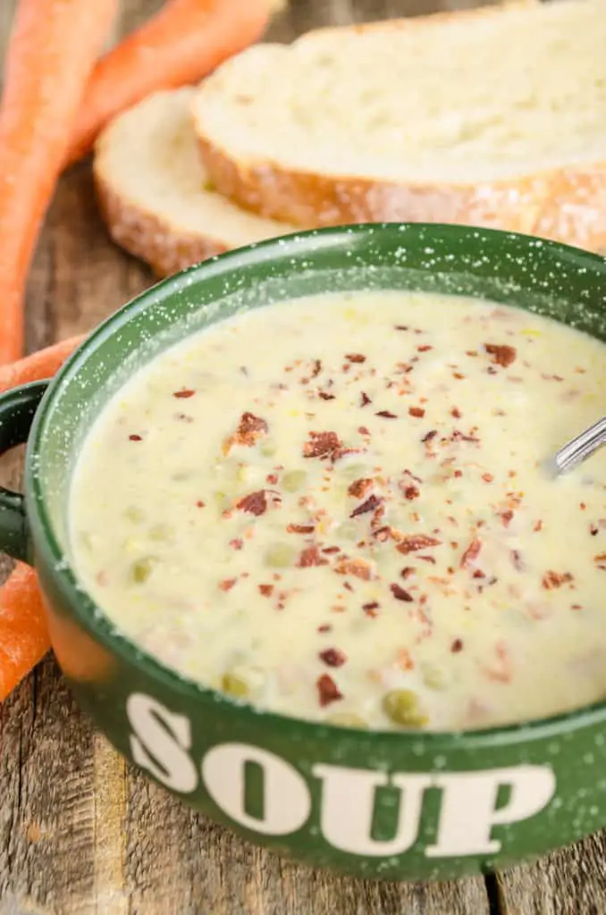 A bowl of Creamy Split Pea and Ham Soup sits surrounded by whole carrots and crusty bread slices. - The Goldilocks Kitchen