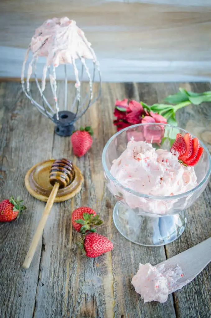 Whipped Strawberry Honey Butter sits in a glass bowl surrounded by fresh strawberries, a honey wand, and a stand mixer whip attachment which also has Whipped Strawberry Honey Butter on it. - The Goldilocks Kitchen