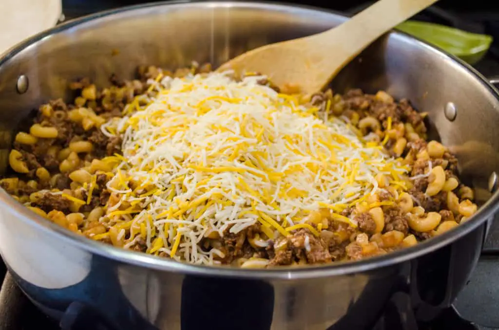 Shredded Mexican cheese blend is added to the beef-macaroni mixture in a skillet to make 30 Minute Chili Mac - The Goldilocks Kitchen