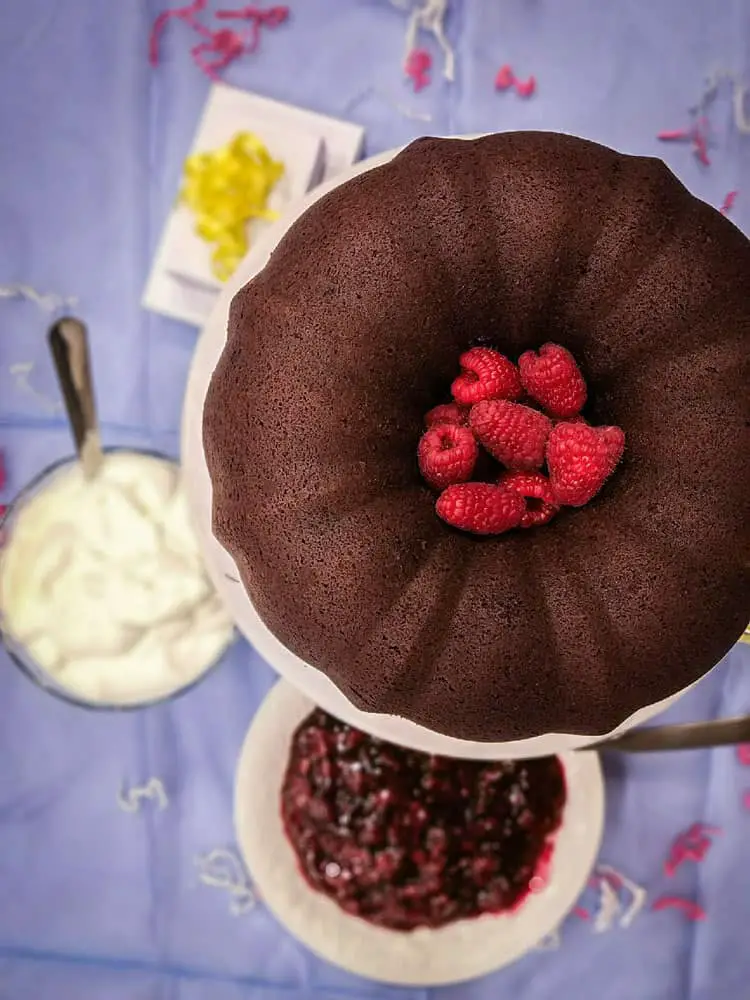A Chocolate Raspberry Bundt Cake in its entirety sits on a cake stand above a bowl of whipped cream and another bowl of raspberry filling. - The Goldilocks Kitchen