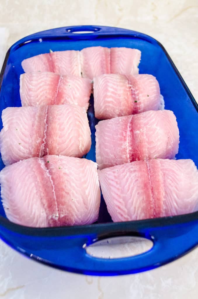 Raw Swai fish bundles stuffed with spinach parmesan stuffing sit in a blue transparent glass baking dish ready to become Creamy Stuffed Fish Florentine - The Goldilocks Kitchen