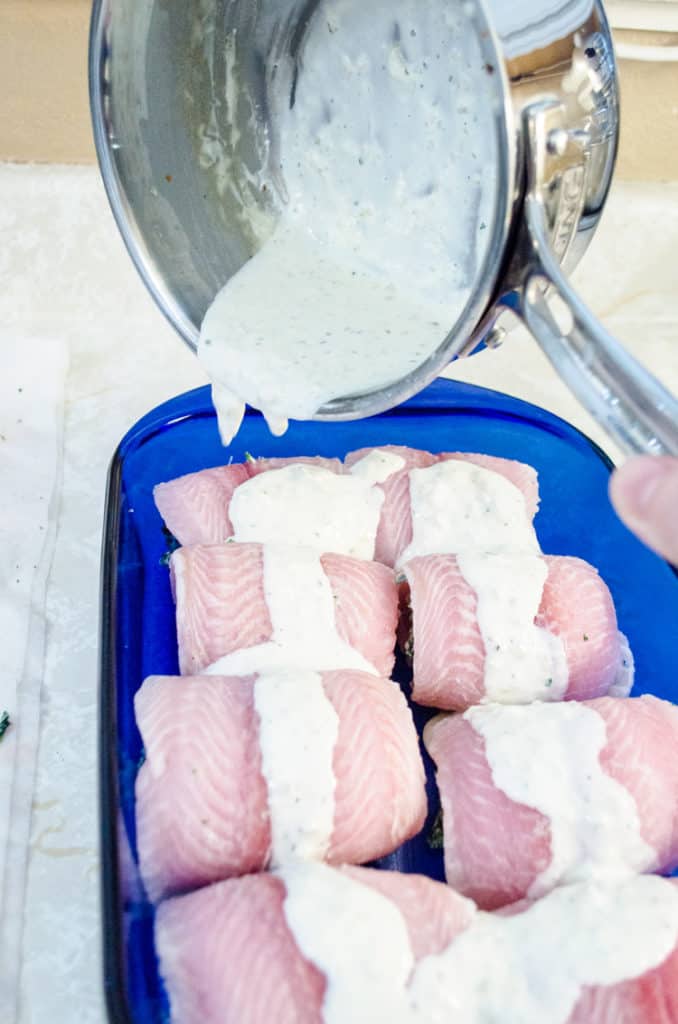 A creamy seasoned white sauce is poured over raw fish bundles that will be baked into Creamy Stuffed Fish Florentine - The Goldilock Kitchen