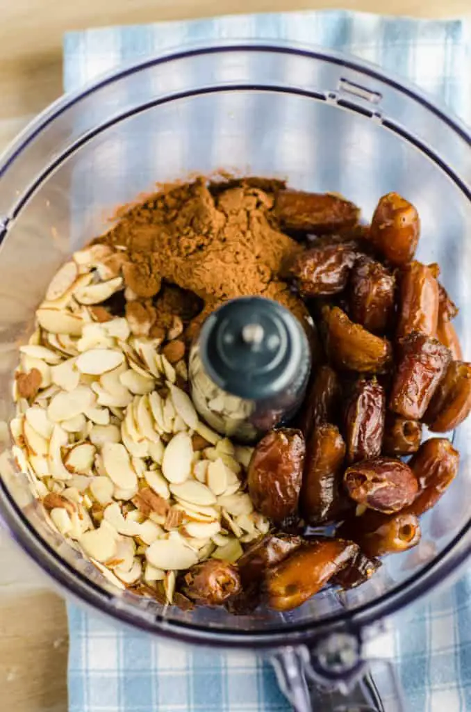 The bowl of a food processor is filled with sliced almonds, pitted whole dates, and cocoa powder for Chocolate Mint Energy Bites - The Goldilocks Kitchen