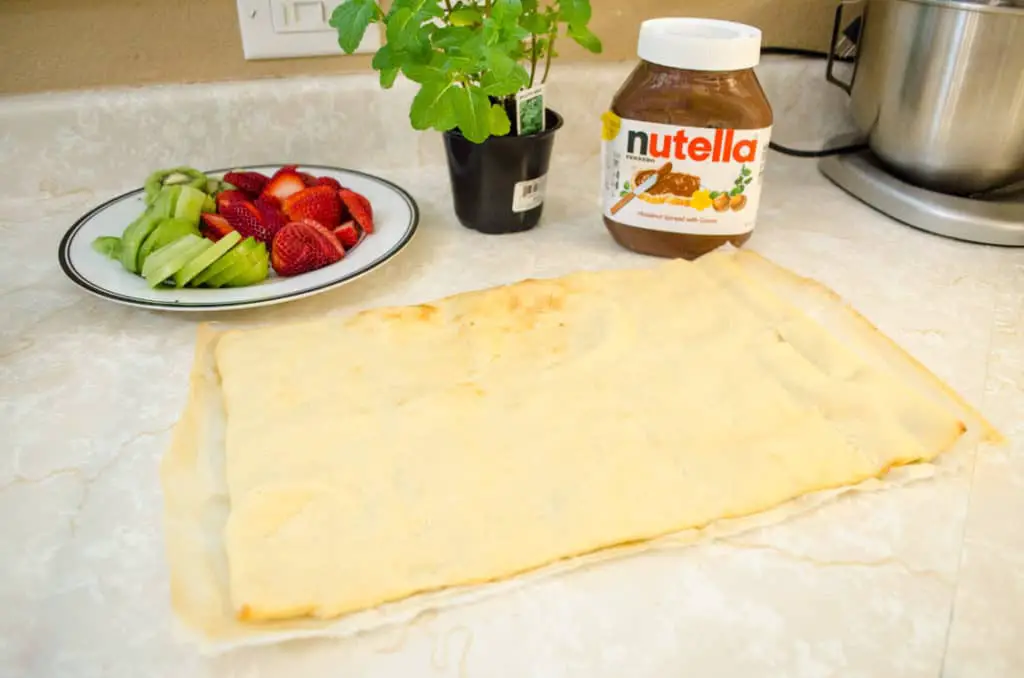 A plate of freshly sliced strawberries and kiwi's, a jar of Nutella and a rectangular cooked pizza crust sit on the counter ready to be turned into Nutella Pizza - The Goldilocks Kitchen
