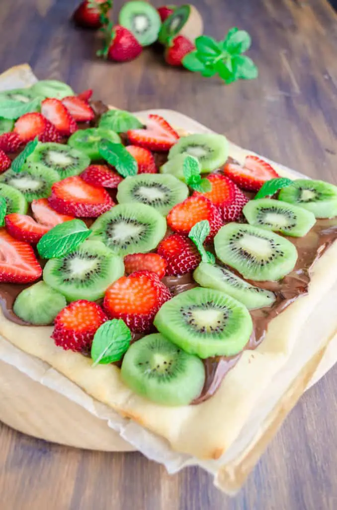 A Nutella Pizza sits on a wooden pizza peel, topped with slices of fresh strawberries and Kiwi with mint leaves sprinkled in here and there - The Goldilocks Kitchen