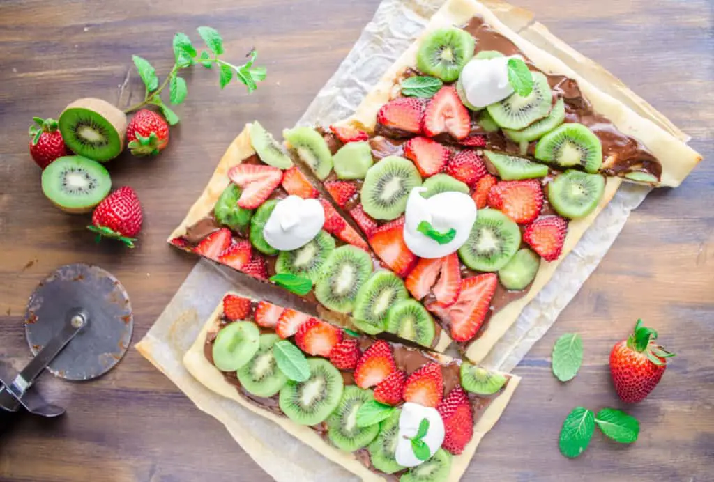 Looking down on a Nutella Pizza sliced into diagonal shapes, surrounded by whole kiwis and strawberries and sprigs of mint - The Goldilocks Kitchen