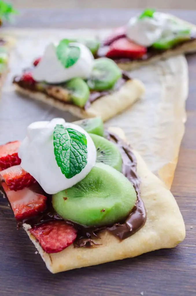 A slice of Nutella Pizza with sliced strawberries and kiwi's sits with a dollop of fresh whipped cream and a mint leaf on top for a garnish - The Goldilocks Kichen