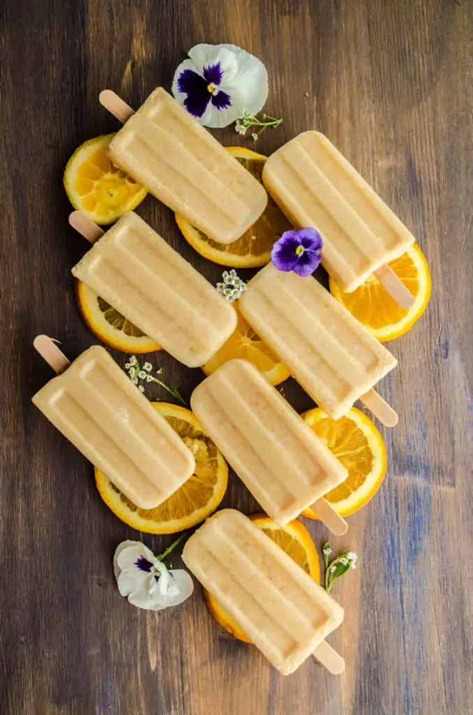 A view looking down on seven Orange Creamsicle Smoothie Pops arranged diagonally, sitting on top of orange slices and surrounded with a few flowers - The Goldilocks Kitchen