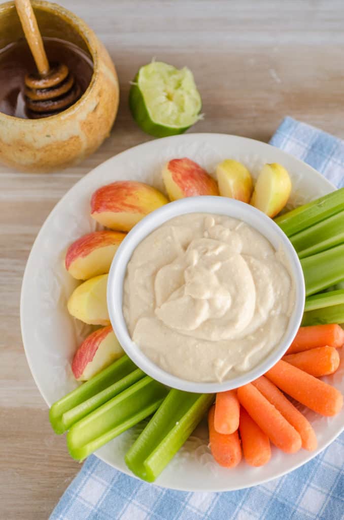 A bowl of Silky Peanut Butter Dip is surrounded with fruits and veggies, a honey pot and a squeezed lime. - The Goldilocks Kitchen