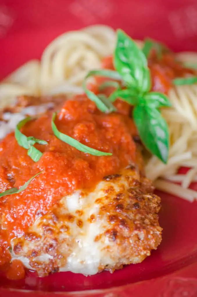 A closeup of Easy Chicken Parmesan on a red plate with a side of spaghetti and some fresh basil for garnish.