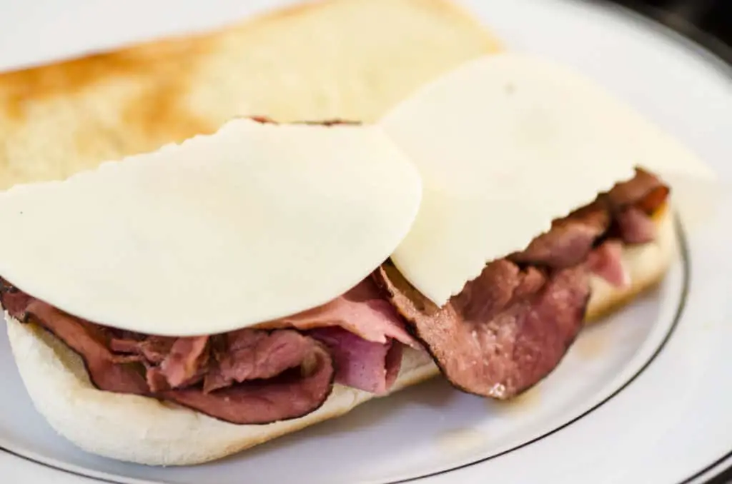 A French Dip with Au Jus sandwich is topped with two torn pieces of provolone cheese that is waiting to be melted.