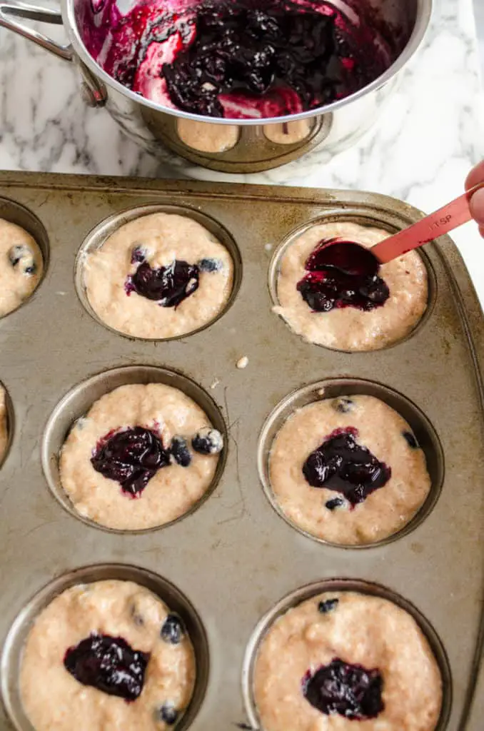 A spoonful of freshly made blueberry jam is placed in the center of each muffin cup full of Moist Whole Wheat Blueberry Swirl Muffins batter.