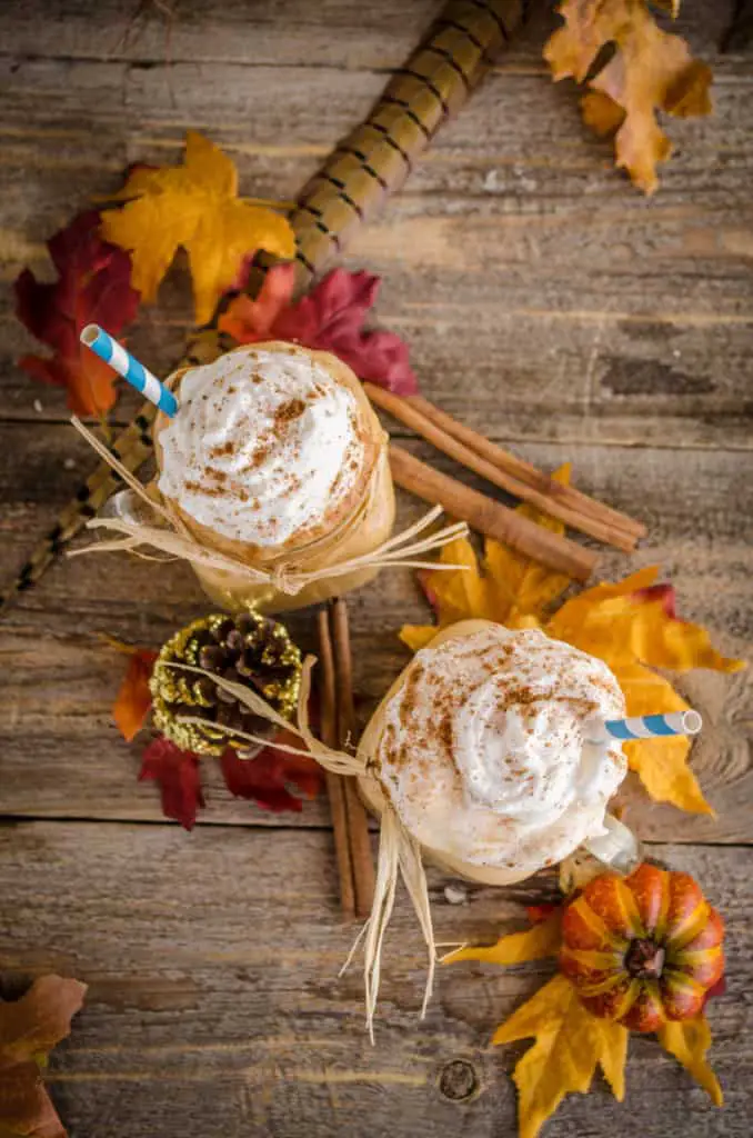 Looking down on a barnwood surface, two Frosty Pumpkin Pie Smoothies with blue striped straws sits in a pile of fall colored leaves.