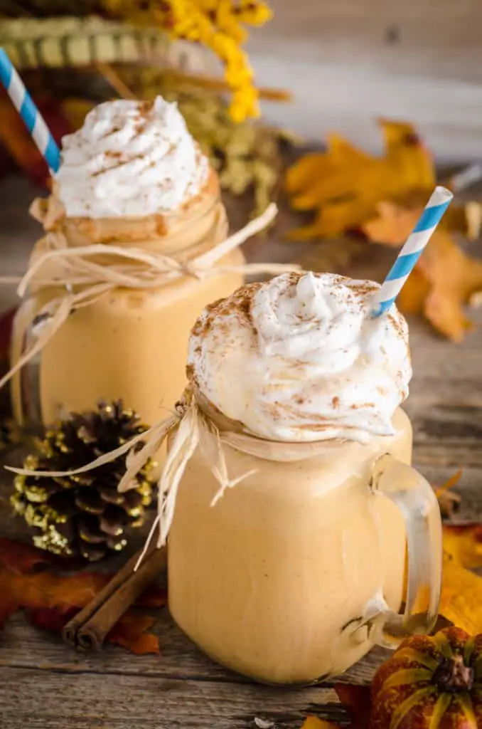 Two Frosty Pumpkin Pie Smoothies sit on a barnwood table surrounded with colorful leaves, shrubbery and a pinecone.