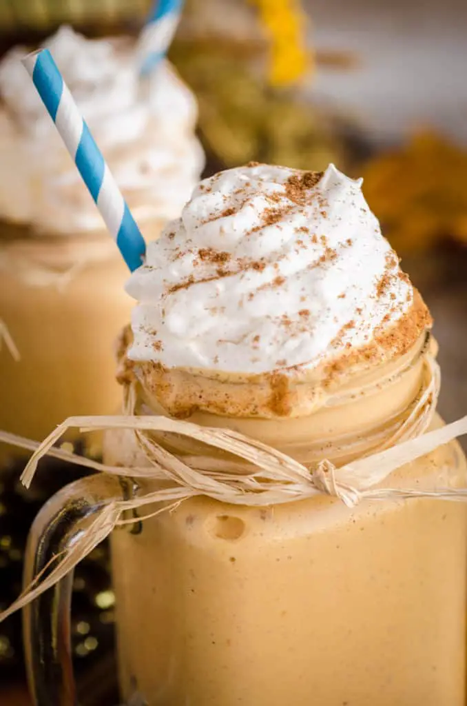 A Frosty Pumpkin Pie Smoothie sits in a glass mason jar mug wrapped with a raffia bow accent, whipped cream and a blue striped straw.