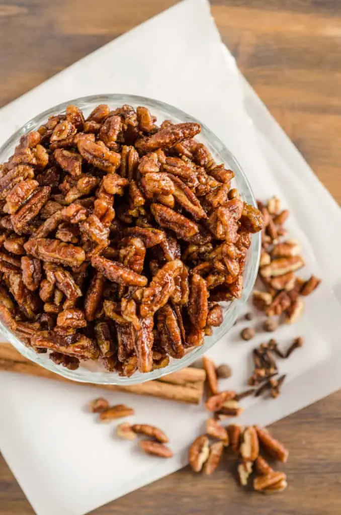 Candied Spice Pecans sit in a glass bowl over parchment paper and a wooden table top - The Goldilocks Kitchen