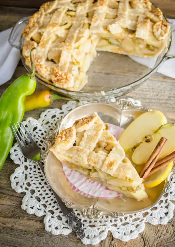 A slice of Green Chile Apple Pie is served up on a silver platter, with the pie and some freshly picked green chiles in the background - The Goldilocks Kitchen