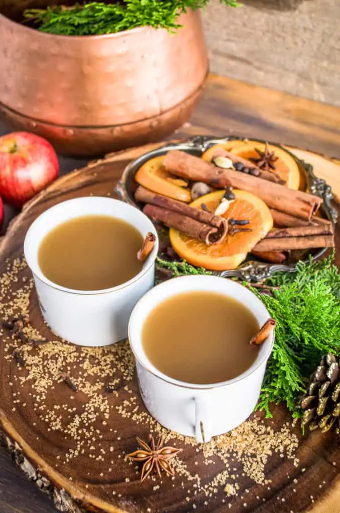 Mugs of Mulled Apple Cider a.k.a. Wassail sit on a wooden table surrounded by sugar, mulling spices and orange slices.