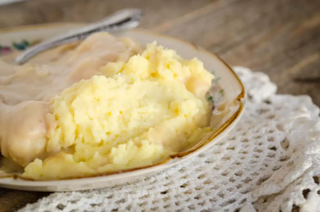 The Best Mashed Potatoes sit on a plate half smothered in gravy - The Goldilocks Kitchen