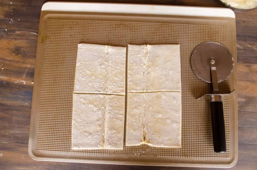 Store bought puff pastry sheets cut into four equal sized squares sit on a baking sheet next to a pizza cutter to make Easy Apple Turnovers - The Goldilocks Kitchen