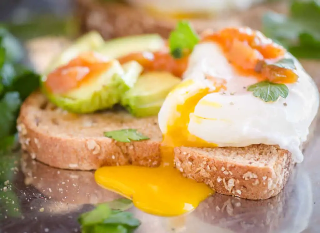 A close up picture of a poached egg spilling it's yolk over the side of a piece of Avocado Toast with Poached Eggs - The Goldilocks Kitchen