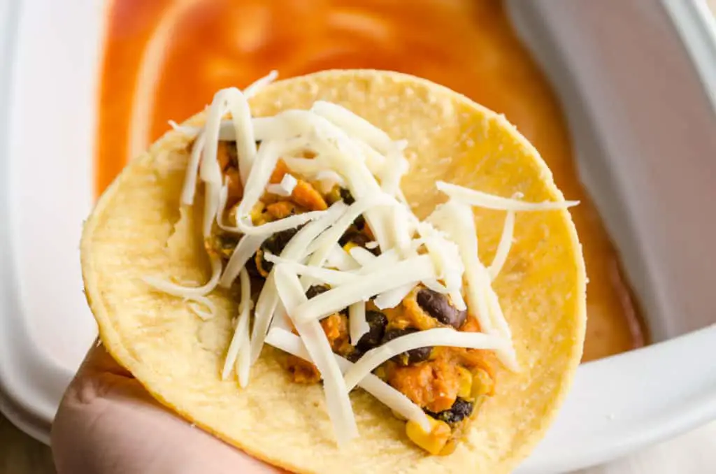 A corn tortilla is held up to show the black bean sweet potato filling sprinkled with cheese to make Black Bean Sweet Potato Enchiladas - The Goldilocks Kitchen