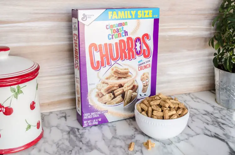 A box of Family Size Cinnamon Toast Crunch Churros Cereal on a countertop, with a white bowl of cereal next to the box for making Churros Snack Mix - The Goldilocks Kitchen