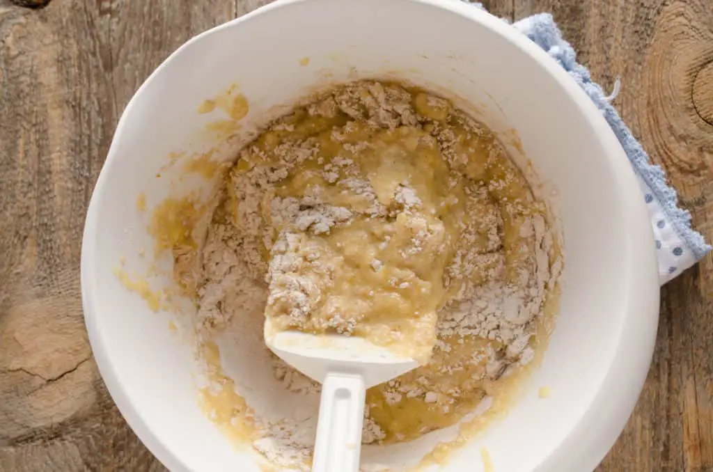 Wet and dry ingredients are gently mixed together in a white mixing bowl with a spatula to make Orange Spice Banana Bread - The Goldilocks Kitchen