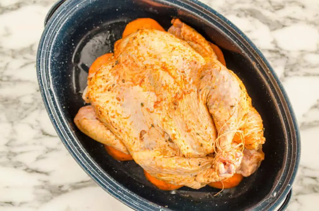 A whole raw chicken rubbed with seasonings and oil is placed over a layer of sweet potatoes in a blue/white speckled enamel oval roasting pan to make Whole Roasted Chicken and Sweet Potatoes - The Goldilocks Kitchen