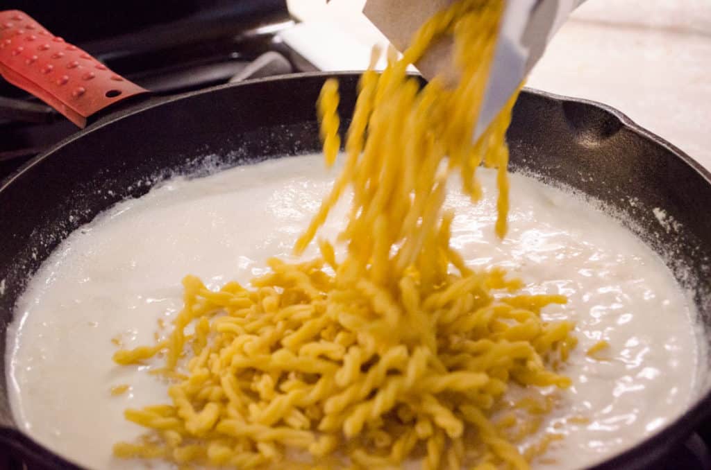 Gemelli pasta is poured into a cast-iron skillet filled with milk to make One-Skillet Bacon Mac and Cheese - The Goldilocks Kitchen