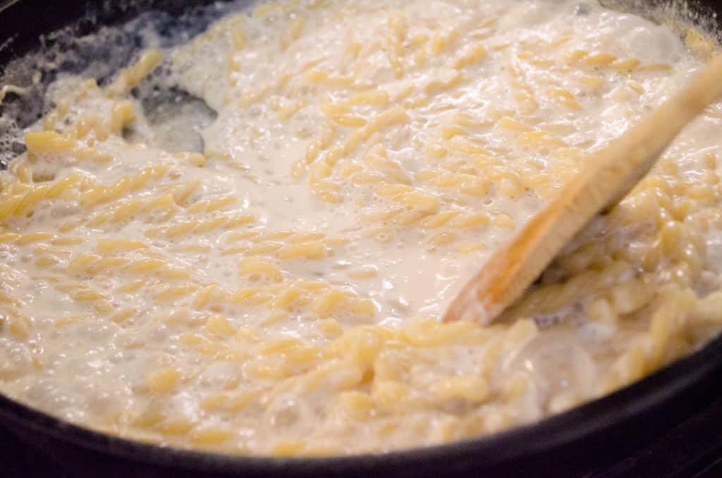 A wooden spoon is scraped along the bottom of a cast-iron skillet filled with cooking pasta in milk and leave a trail. One-Skillet Bacon Mac and Cheese - The Goldilocks Kitchen