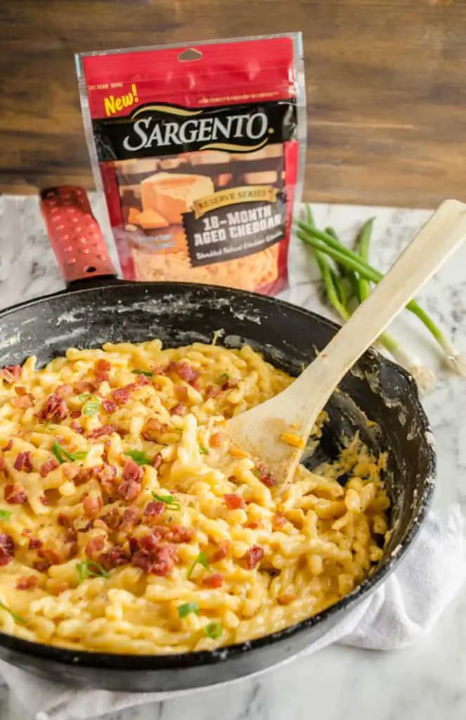 A closeup picture of a bag of Sargento Reserve Series 18-Month Aged Cheddar and a few green scallions, beside a cast iron skillet with One-Skillet Bacon Mac and Cheese in it. - The Goldilocks Kitchen