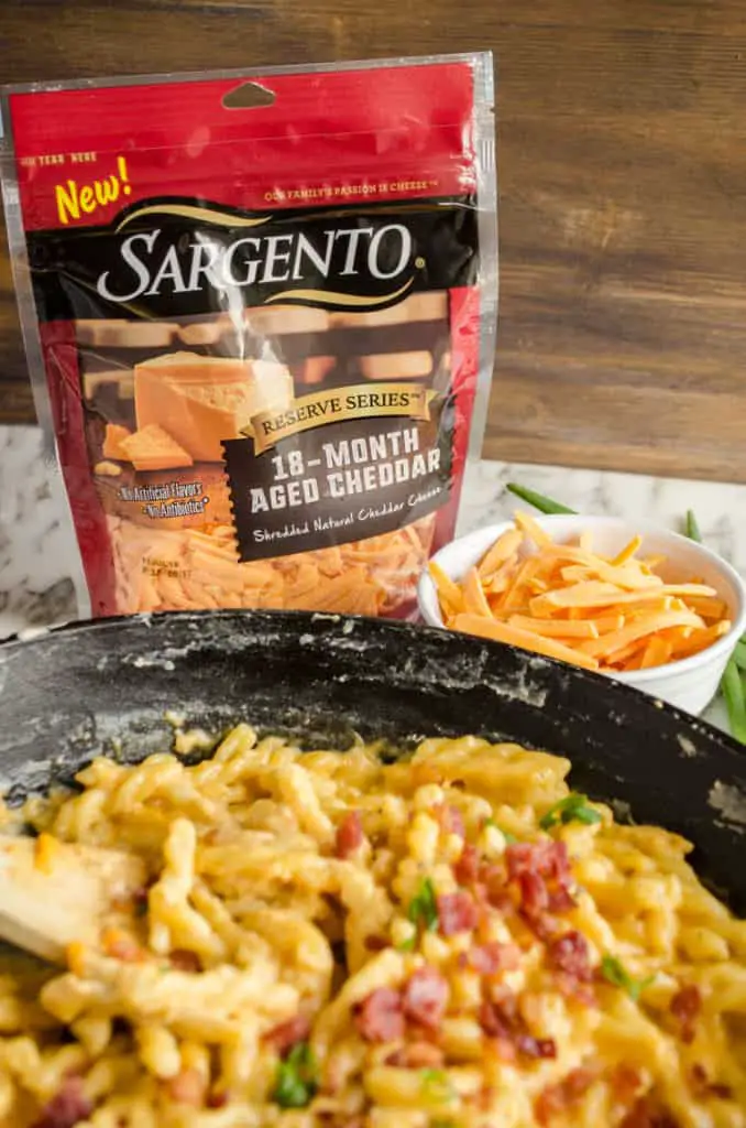 A closeup picture of a bag of Sargento Reserve Series 18-Month Aged Cheddar, beside a bowl of shredded cheddar and a cast iron skillet with One-Skillet Bacon Mac and Cheese in it. - The Goldilocks Kitchen