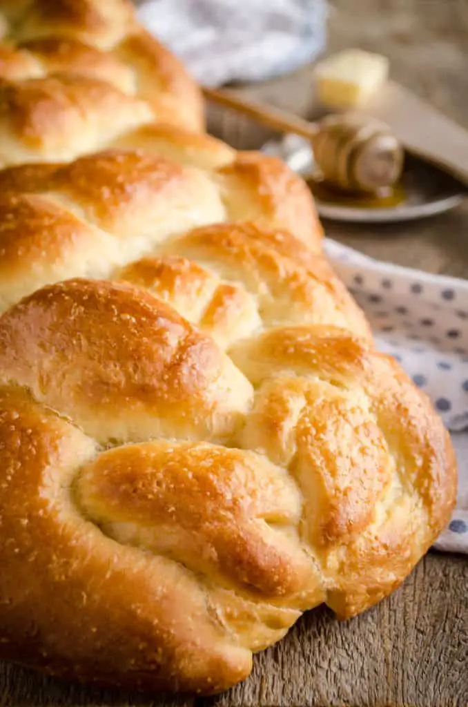 A closeup of freshly baked Braided Challah Bread on a wooden table with a honey wand and a pat of butter - The Goldilocks Kitchen