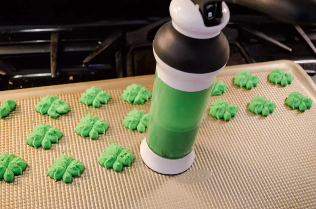 Green clover shaped cookies are pressed out of a cookie press and onto a non-stick cookie sheet to make St. Patricks Day Cream Cheese Spritz Cookies - The Goldilocks Kitchen