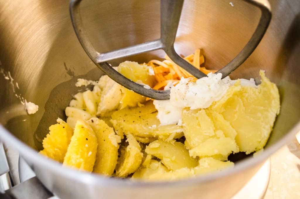 A stand mixer bowl filled with baked potato, sour cream, shredded cheddar cheese, Boursin cheese, melted butter and salt and pepper for Make Ahead Twice Baked Potatoes - The Goldilocks Kitchen