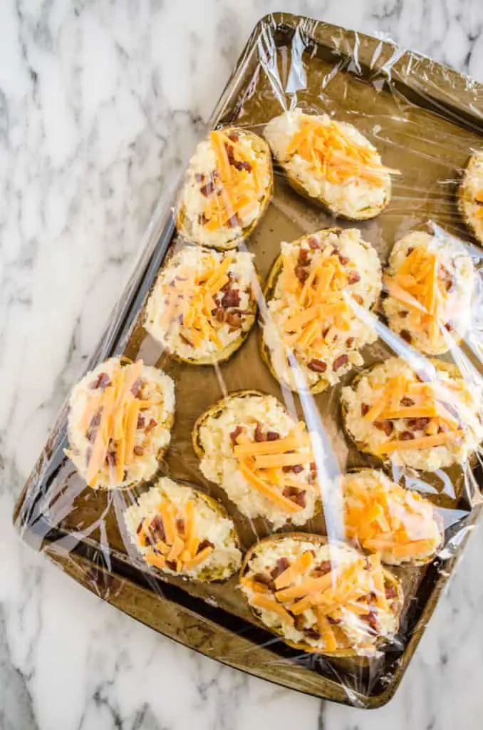 Prepared Make Ahead Twice Baked Potatoes sit on a rimmed baking sheet covered with plastic wrap - The Goldilocks Kitchen