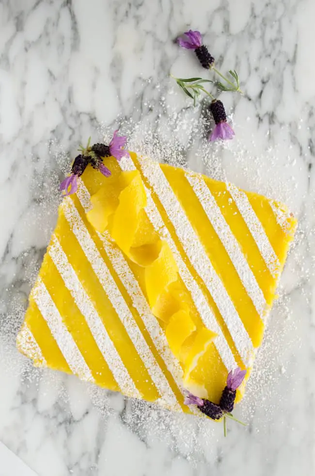 A batch of Goldilocks Kitchen Lemon Bars garnished with stripes of confectioner's sugar, a lemon peel spiral in the center and purple lavender flowers at the corners -The Goldilocks Kitchen
